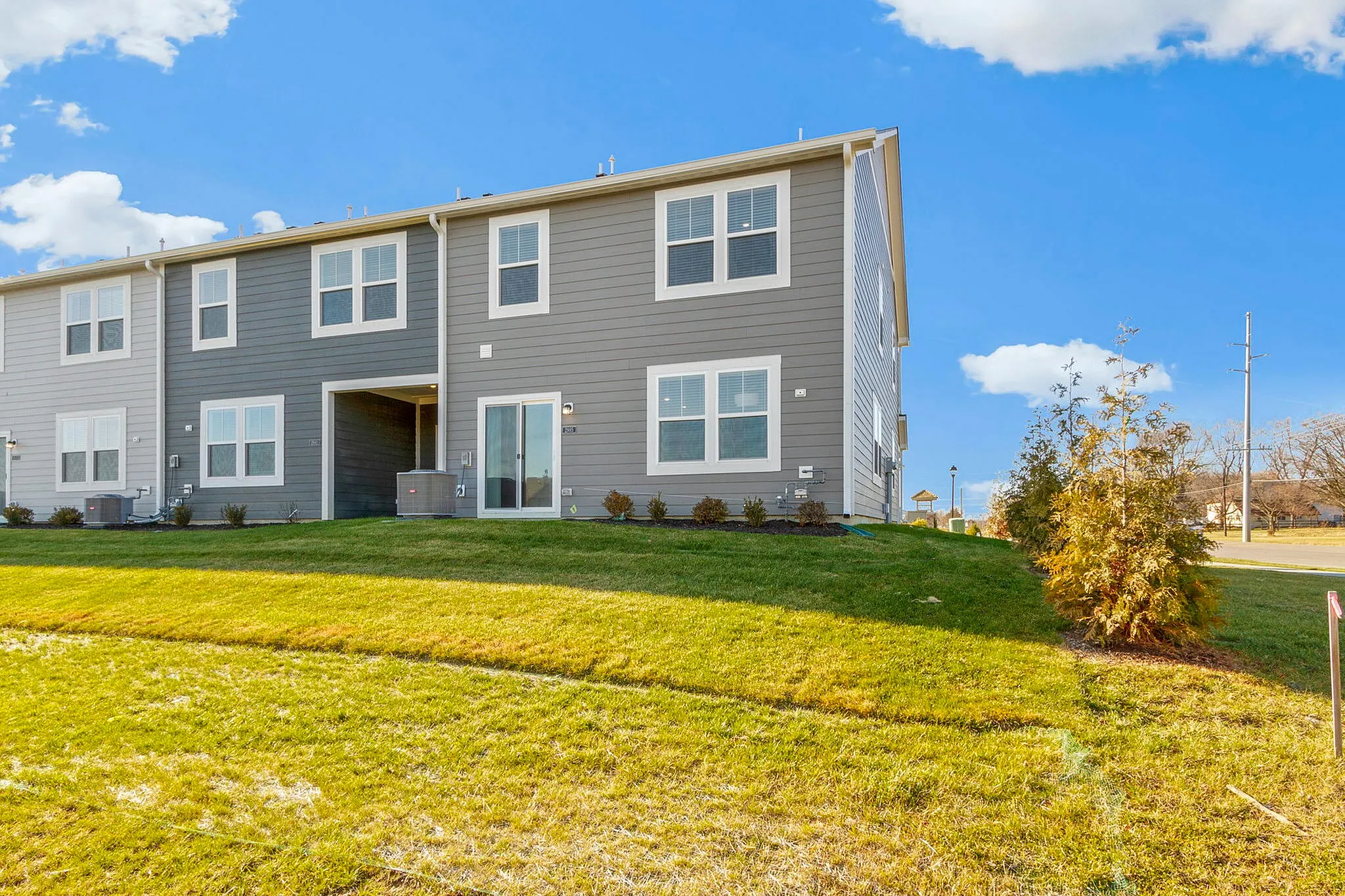 Townhomes in Brownsburg, IN - Wynne Farms - Rear Townhome Exterior With Gray Siding, White Trim, Large Windows, Patio Doors, Green Lawn, Landscaping, And A Bright Blue Sky.