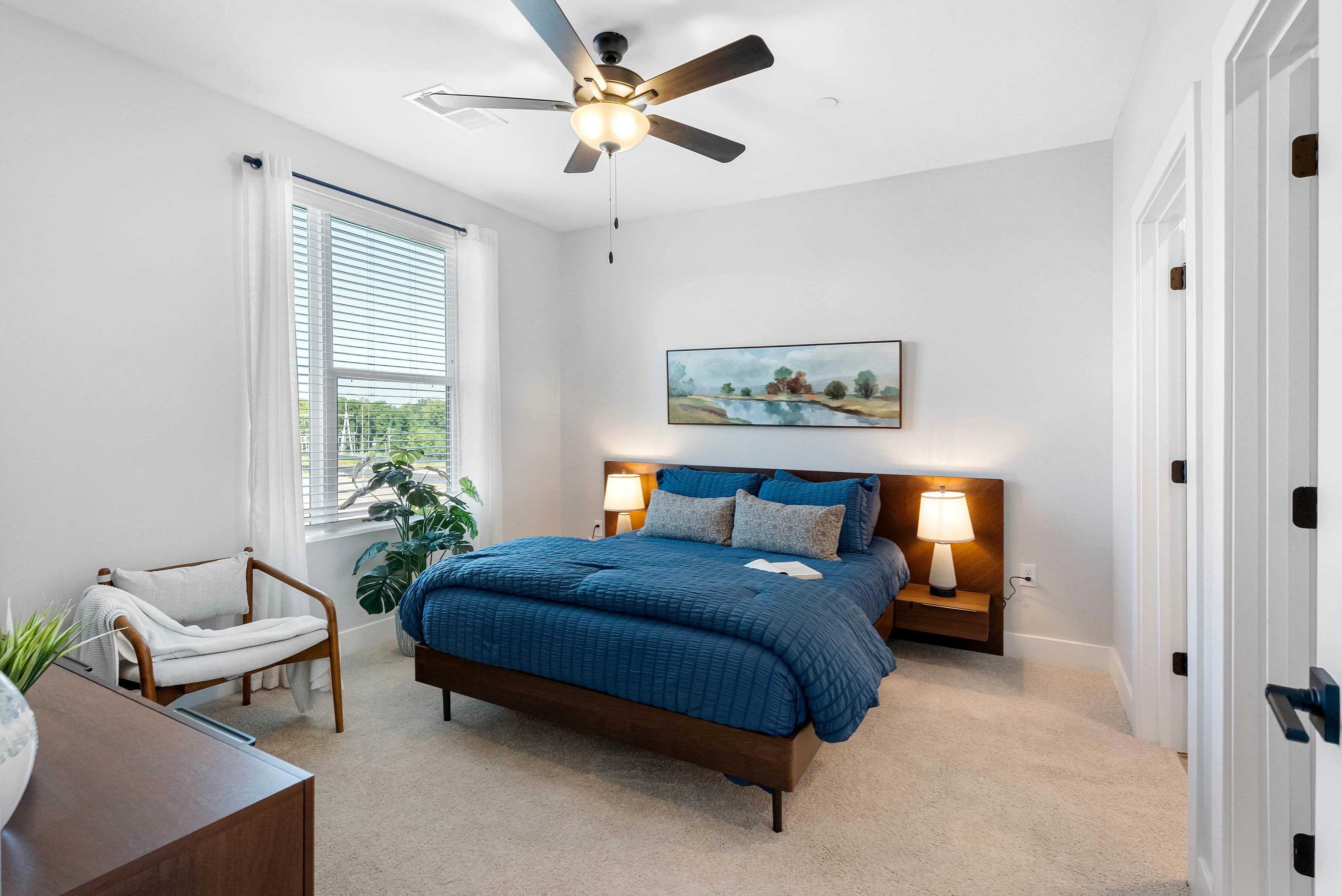 North Chesterfield, VA, Pet-Friendly Apartments - The James at Springline - A Bedroom with Nightstands, Lamps, Ceiling Fan, Large Window, Dresser, and Indoor Plant.