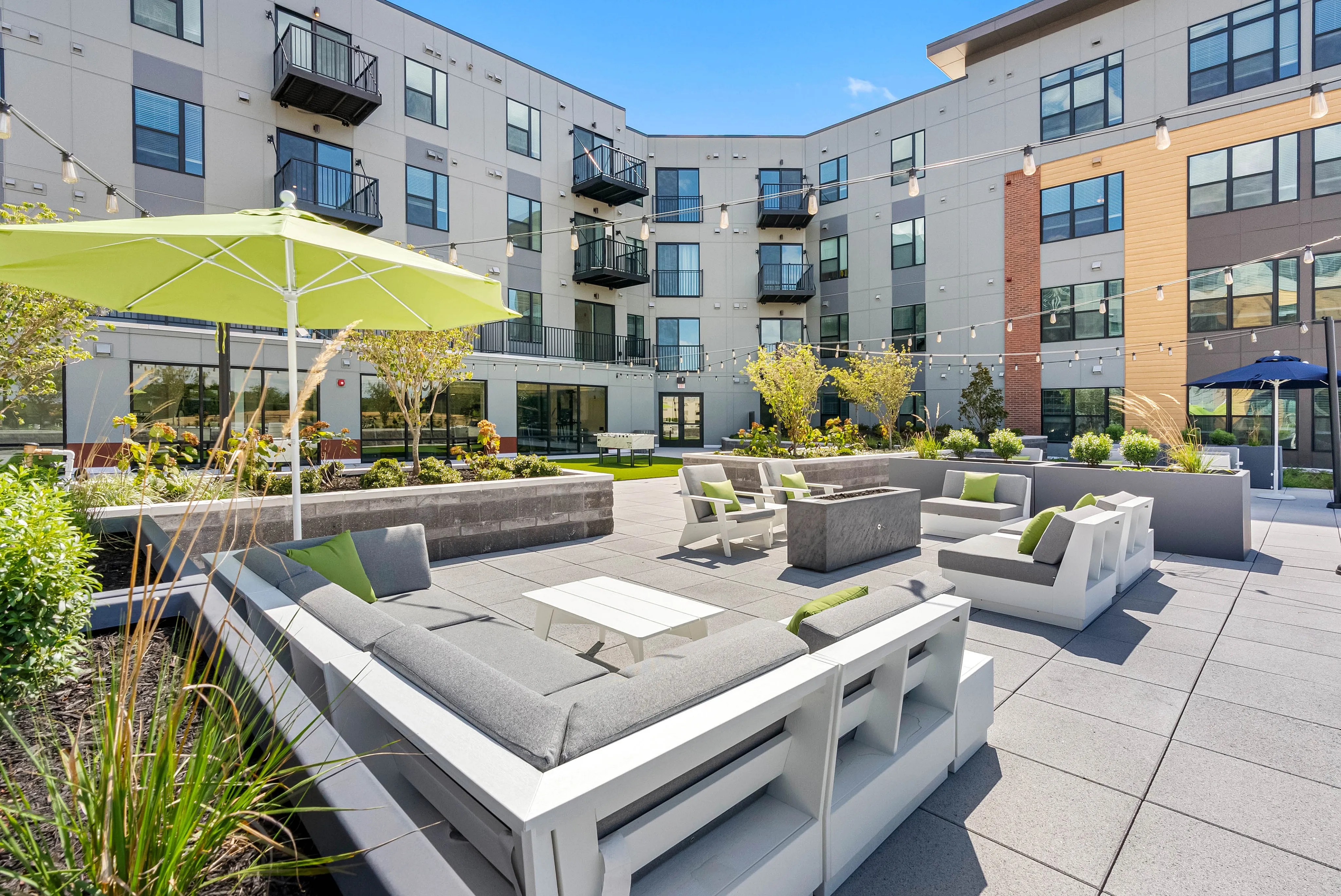 High-End Apartments in Richmond, VA - The James at Springline - An Outdoor Courtyard with Cushioned Lounge Seating, Patio Tables, Umbrellas, String Lighting, and Landscaped Planters.