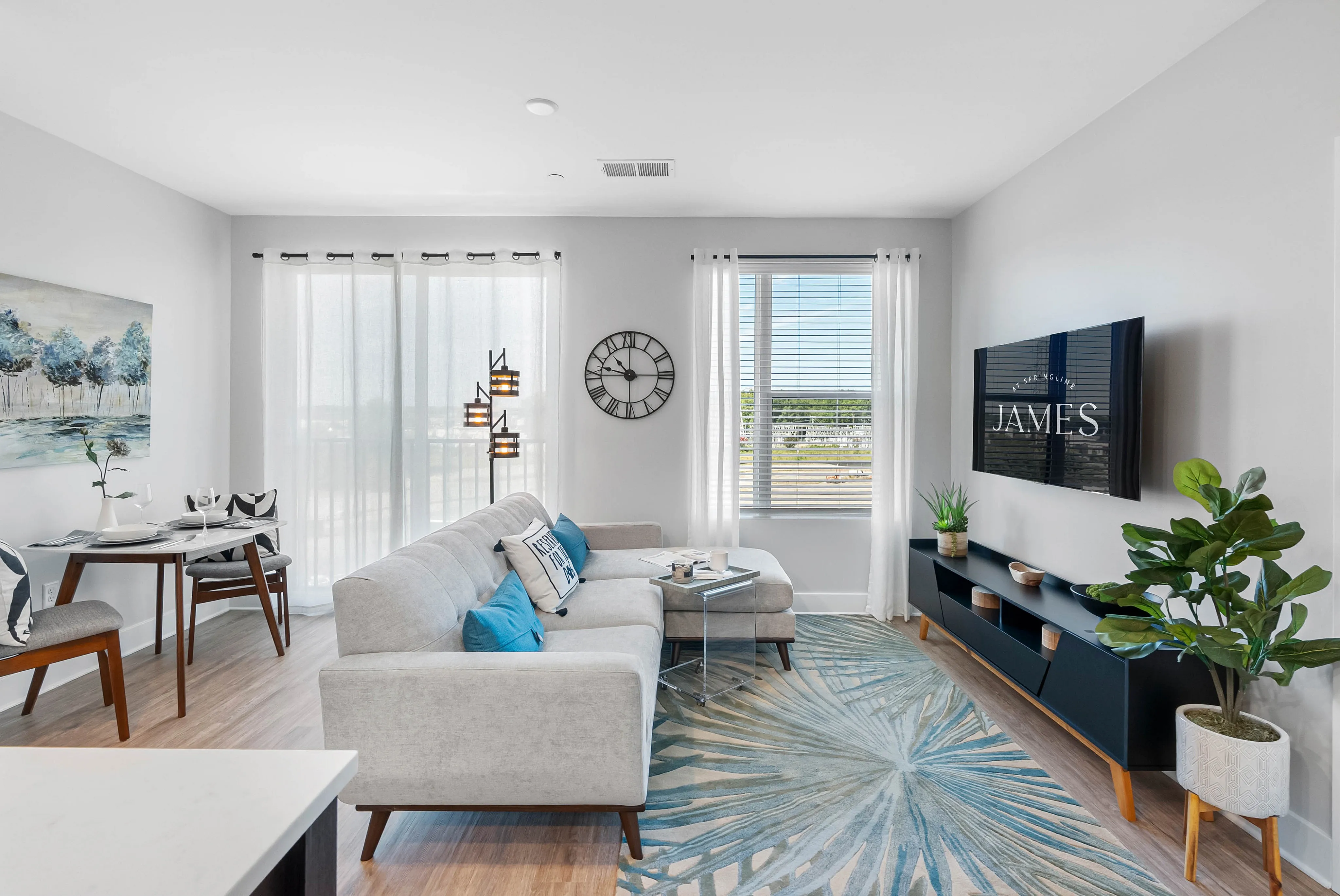 North Chesterfield, VA, Apartments Near Chesterfield Town Center - The James at Springline - A Living Room with a Sectional Sofa, Wall-Mounted TV, Large Windows, and Indoor Plants.