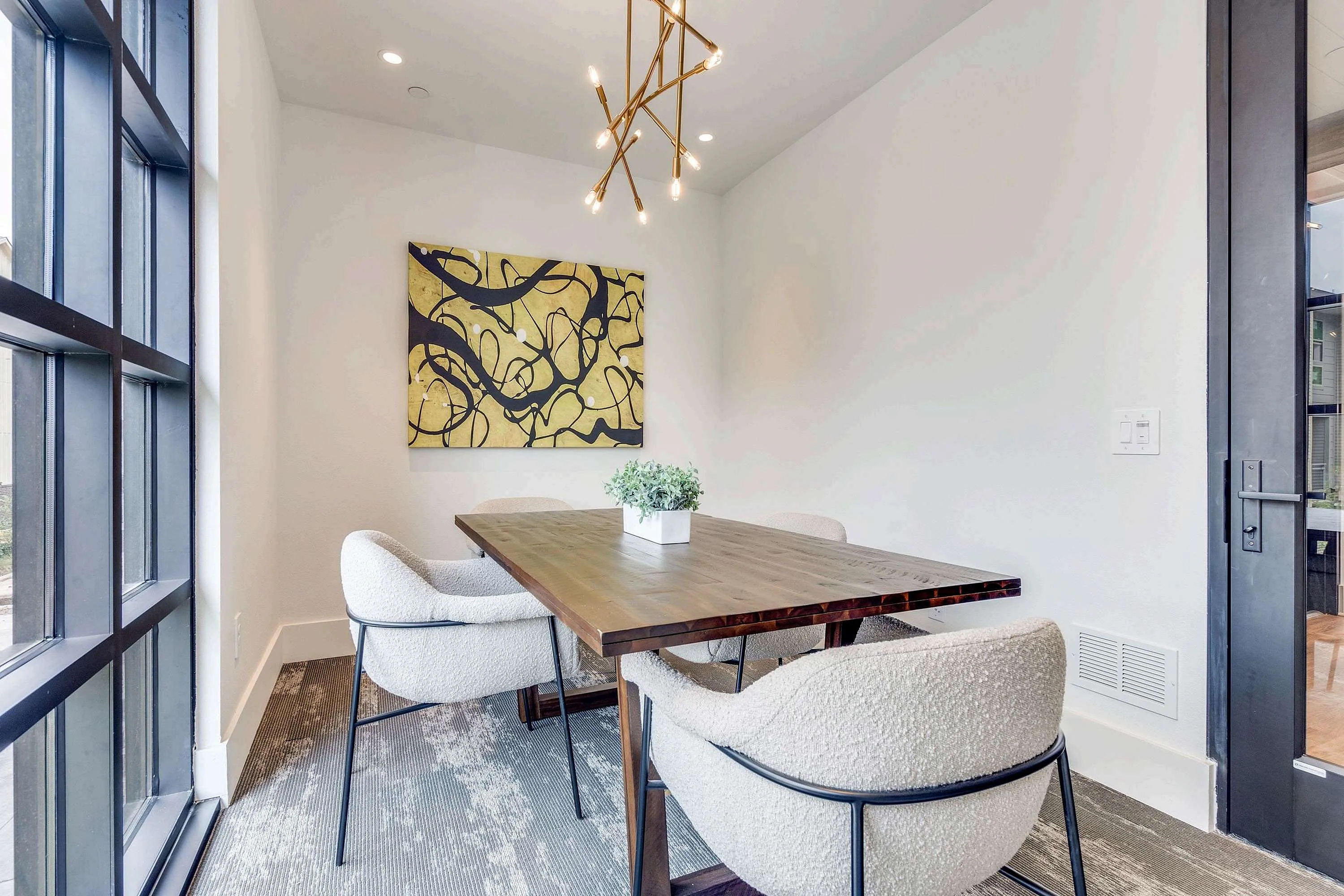 A modern conference area features a wooden table surrounded by two white upholstered chairs, with a vibrant abstract painting on the wall and a small potted plant as a centerpiece. Natural light streams in through large windows, creating a bright and inviting atmosphere.