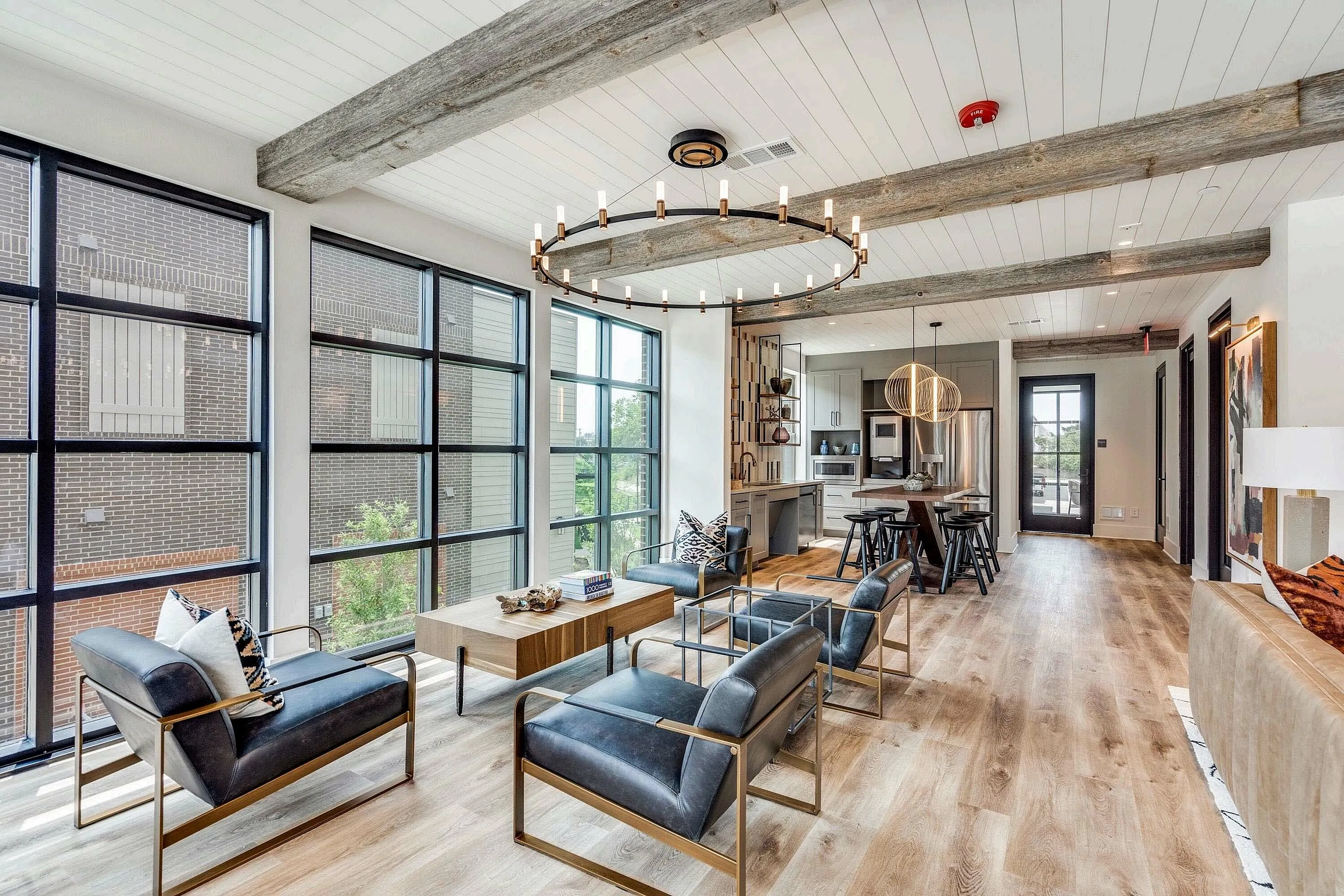 A modern clubhouse space featuring sleek black chairs, a large round chandelier, and large windows that allow natural light. The open layout connects the living area to a kitchen in the background.