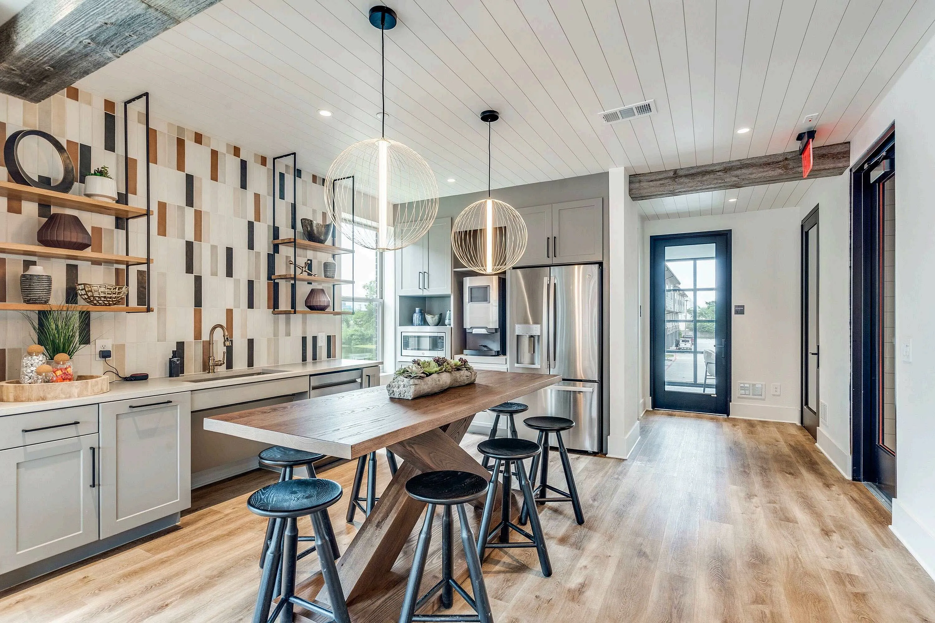 A modern kitchen featuring a wooden dining table with black stools, stylish shelving on the wall, and large windows allowing natural light. The space has a contemporary design with light wood flooring and sleek appliances.