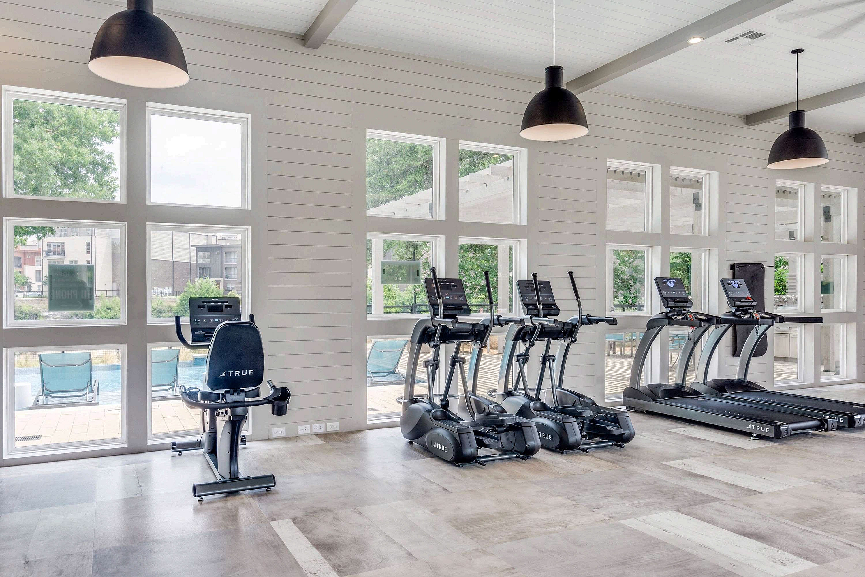 Bright fitness room featuring exercise equipment including treadmills and an elliptical, with large windows providing natural light and a view of the outdoor pool area.