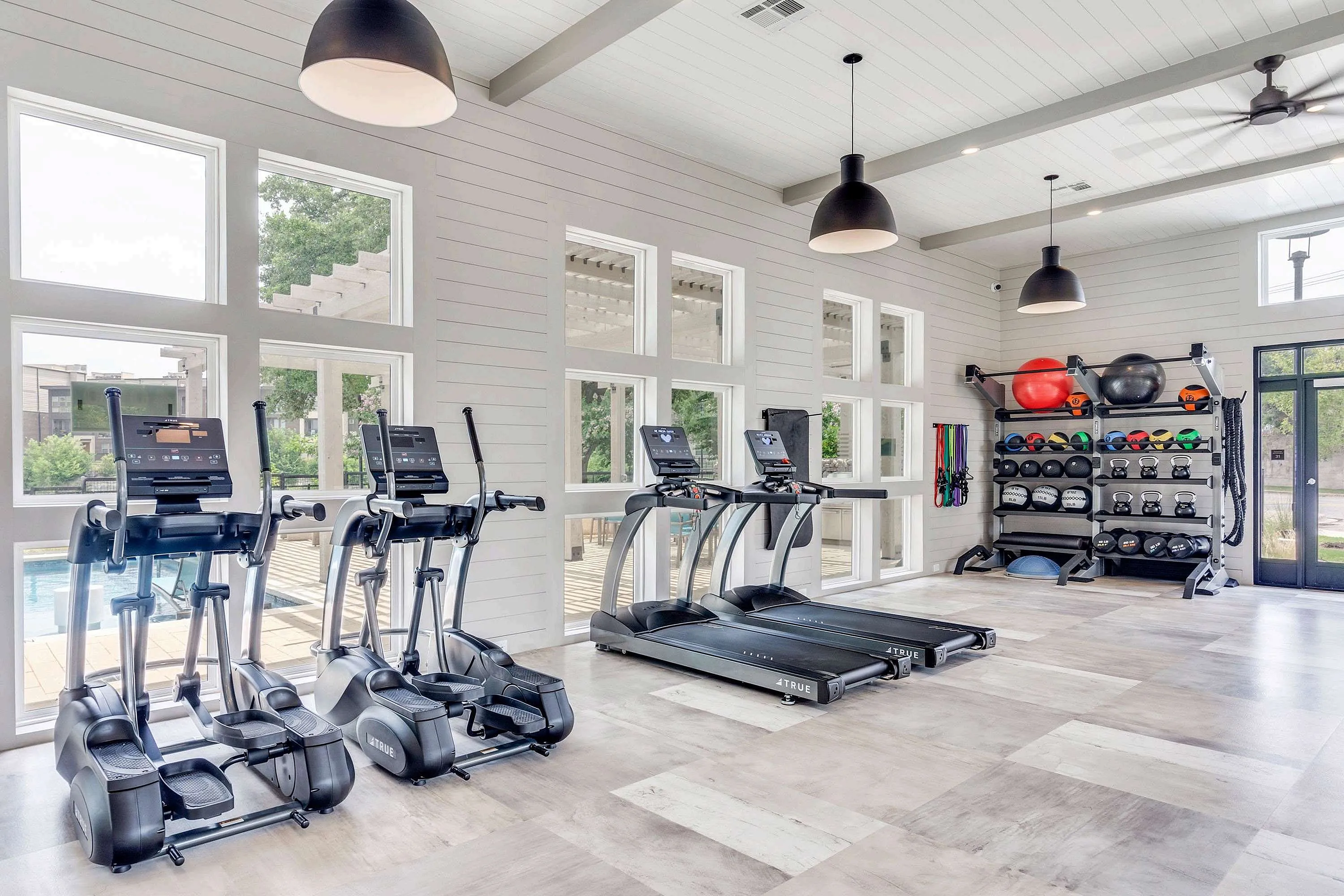 A bright fitness room featuring several treadmills and elliptical machines, with large windows letting in natural light. The space includes a storage area for fitness equipment along one wall.