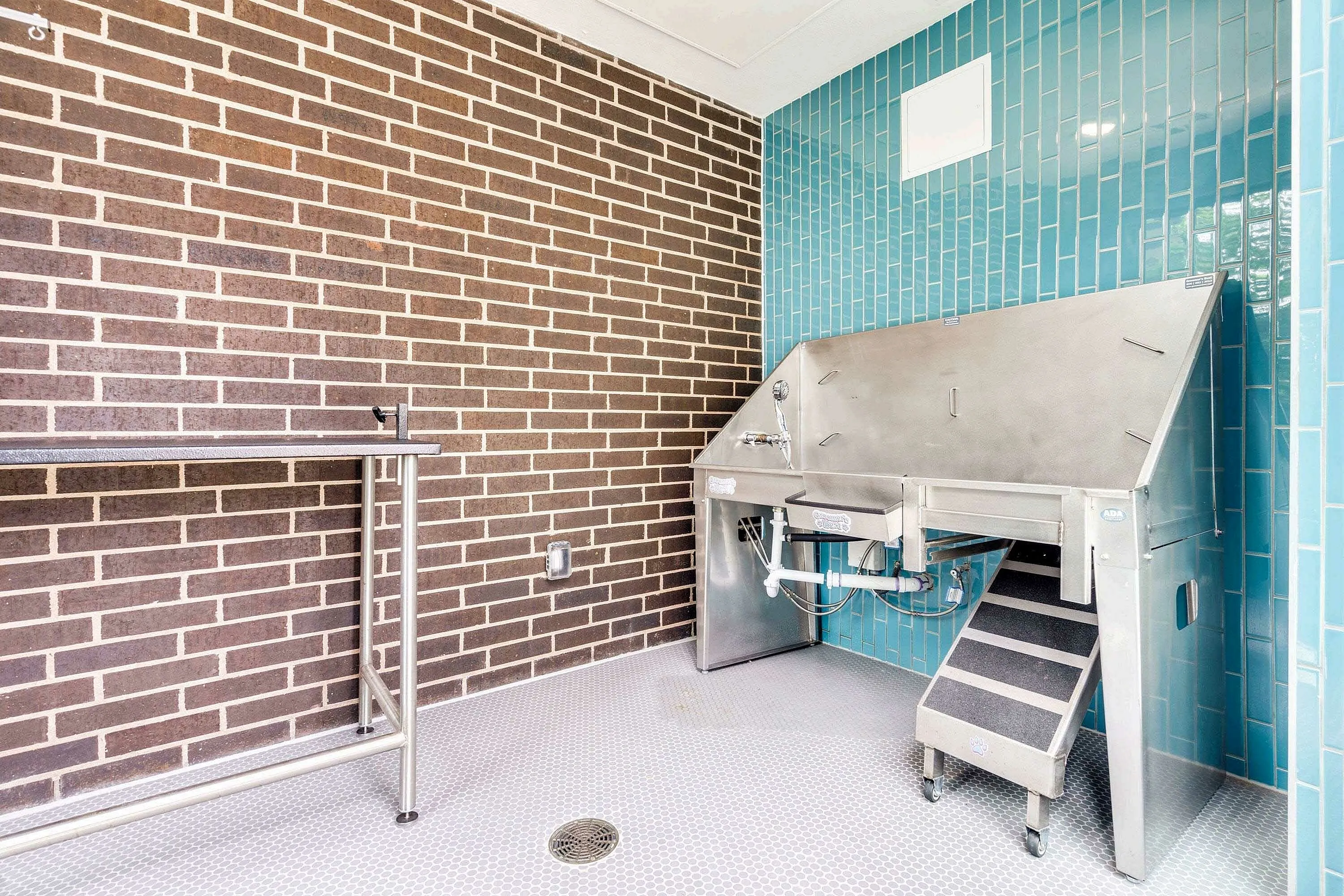 A stainless steel pet grooming station is set against a backdrop of brown brick and turquoise tiles, featuring a set of stairs for easy access. The space is clean and well-lit, designed for pet grooming.