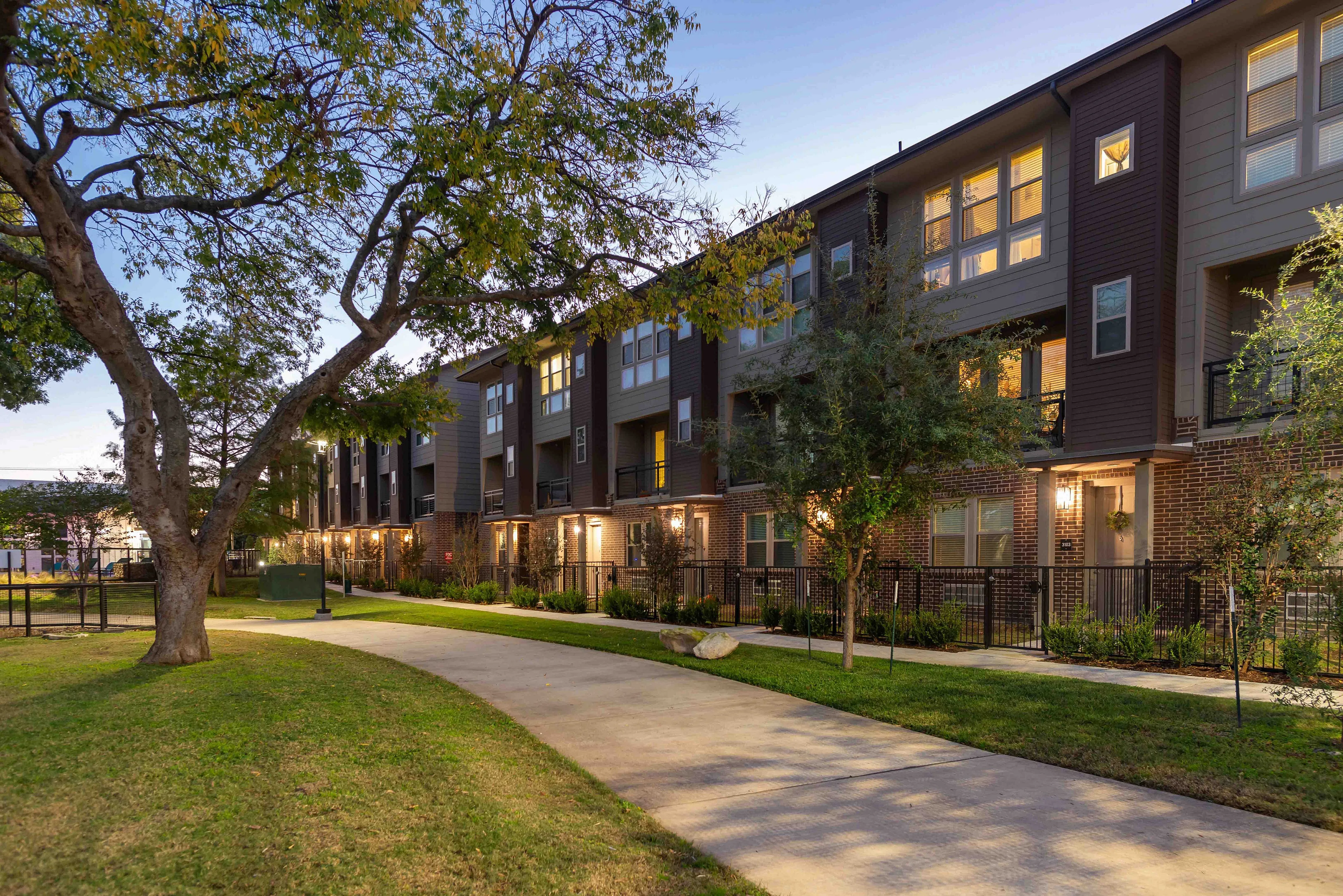 Exterior homes surrounded by trees and a landscaped pathway, is shown during twilight. The warm lights from the windows create a welcoming atmosphere.