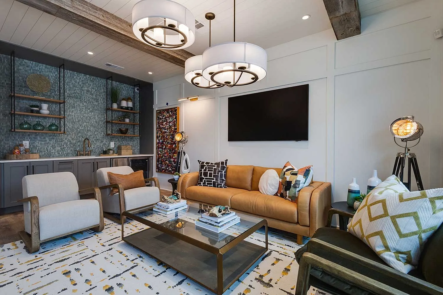 A stylish clubroom featuring a brown leather sofa, two white armchairs, and a coffee table with decorative items. The space is illuminated by modern light fixtures and has a large TV mounted on the wall, with a patterned rug on the floor.