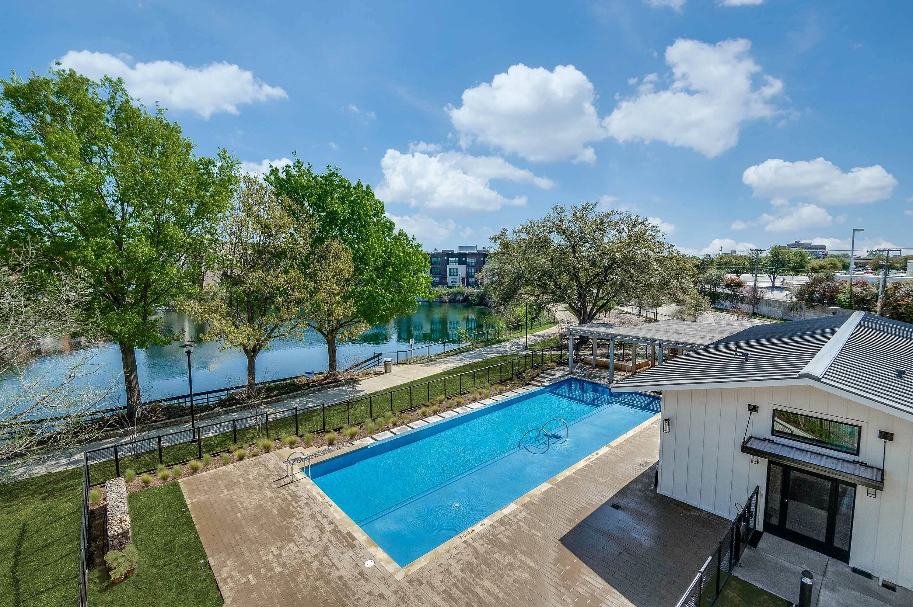 A clear view of a swimming pool surrounded by greenery, with a building nearby and a serene lake in the background under a bright blue sky.