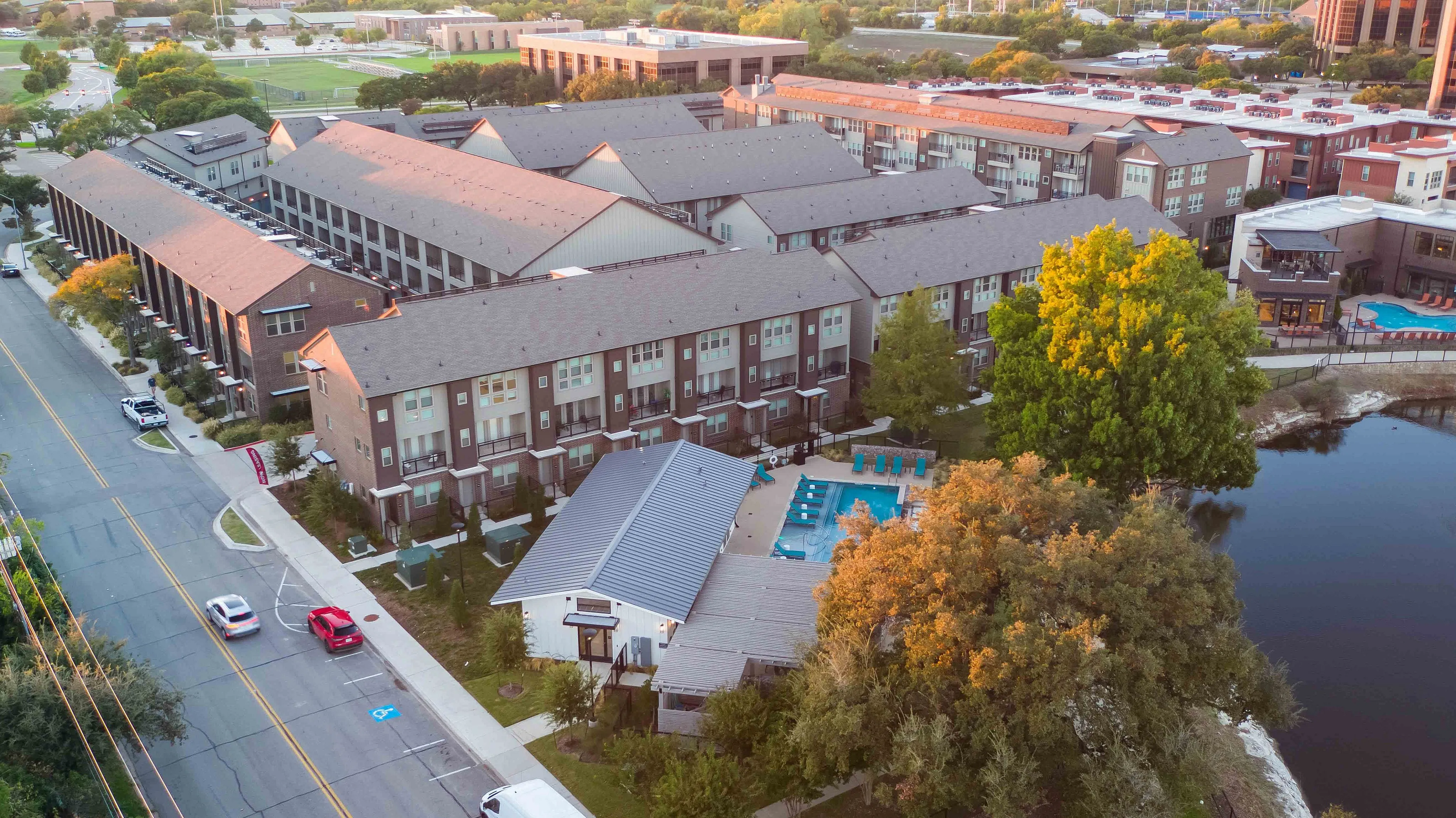 Aerial view of a residential complex with multiple buildings, a swimming pool, and a nearby pond. The image shows a tree-lined street and green spaces in the surrounding area.