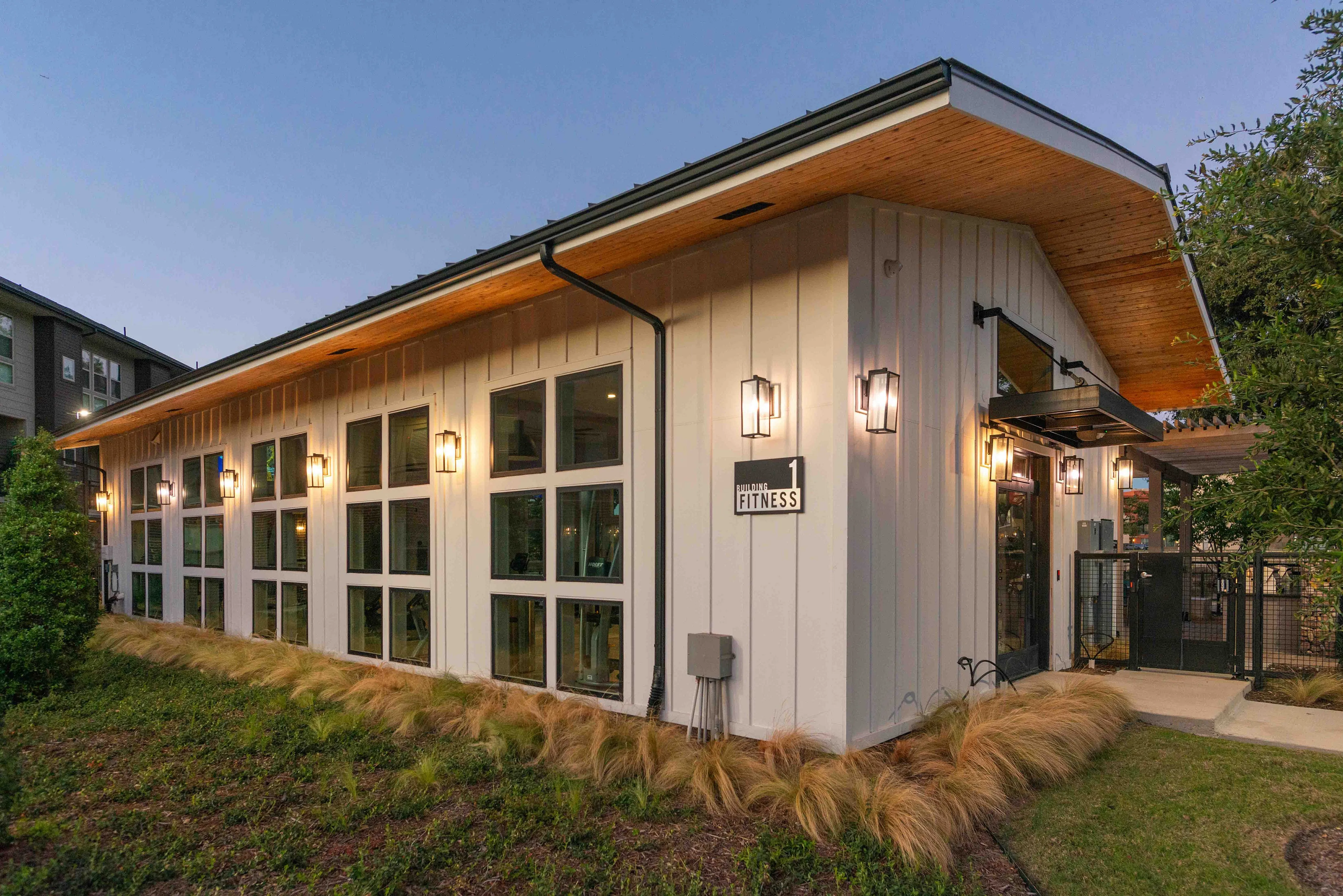 A modern building with large windows and a wooden overhang, surrounded by grassy landscaping. The exterior features a mix of white and natural wood elements, creating a welcoming atmosphere.