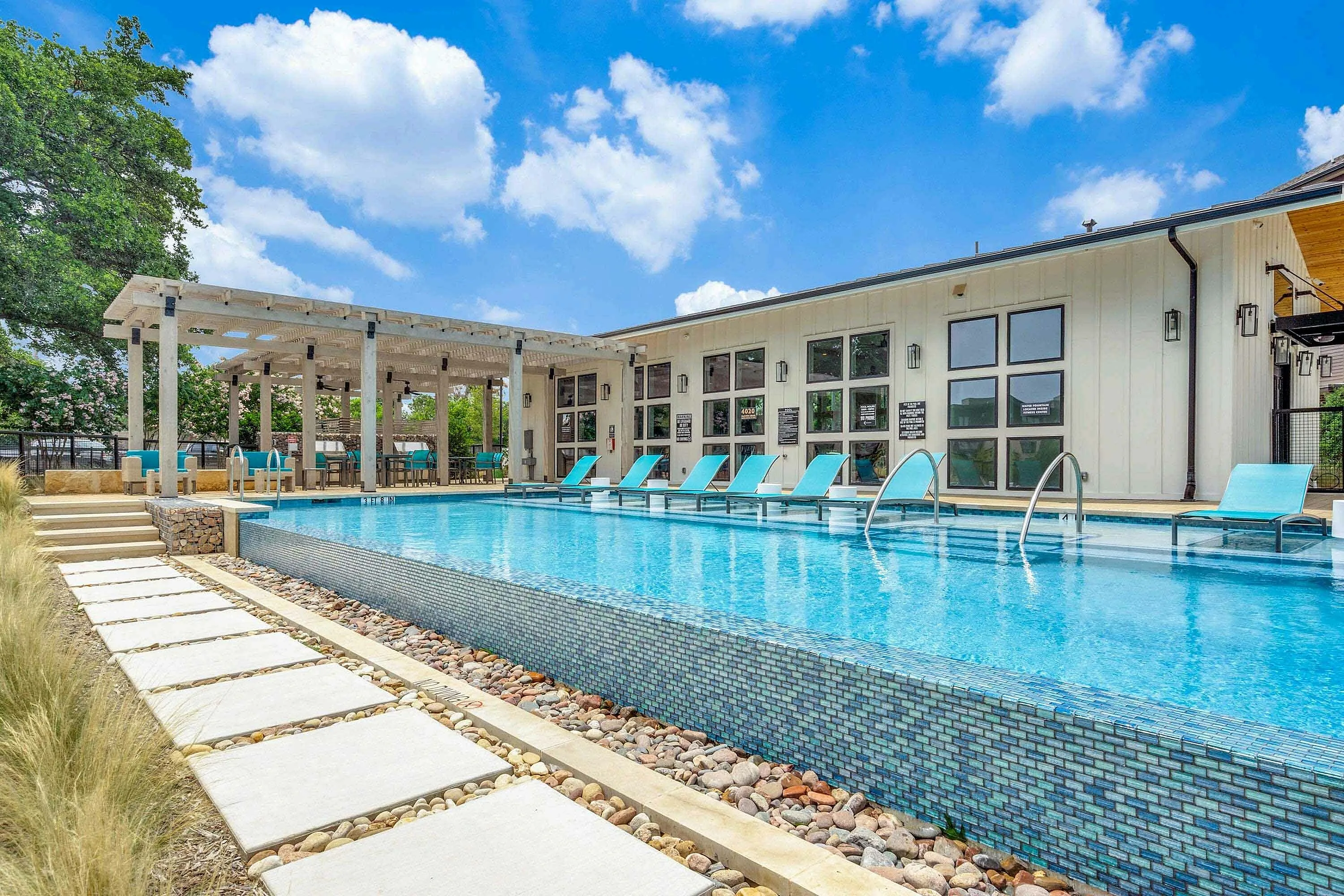 A modern outdoor swimming pool surrounded by lounge chairs and a patio area, set against a bright blue sky with fluffy clouds. The pool area features a sleek design with stone pathways and greenery nearby.