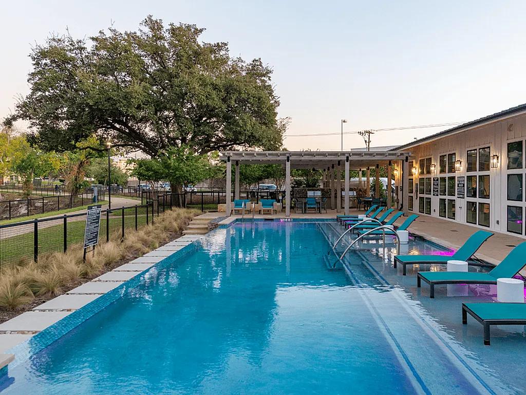 A clear blue swimming pool is surrounded by lounge chairs and a shaded seating area, with trees in the background. The scene captures a relaxing outdoor space, ideal for leisure and socializing.