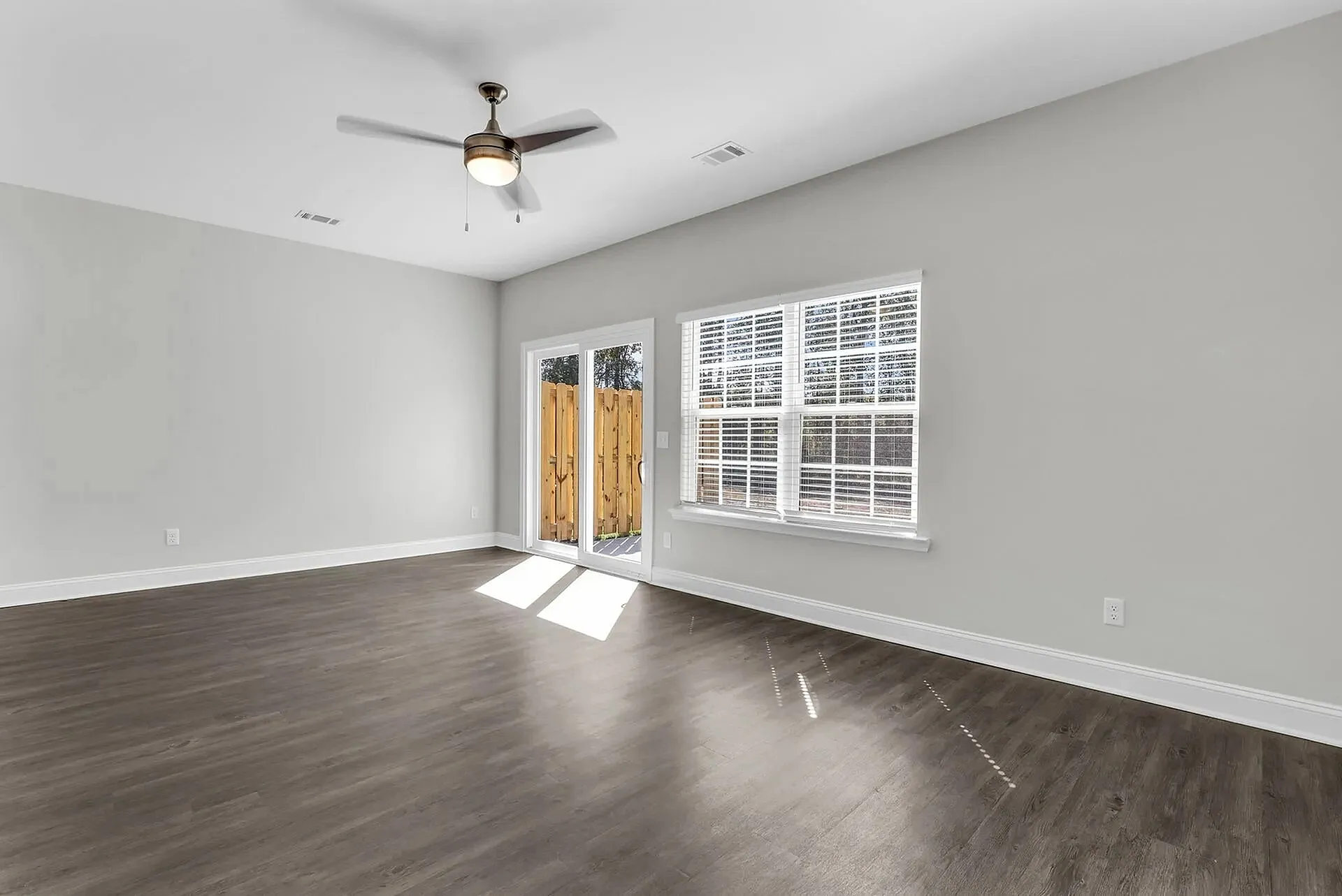 Statesboro, GA, Townhomes with Attached Garage - The Outpost - Spacious Living Room With Wood-Style Flooring, Ceiling Fan, Wide Window, And Glass Patio Door To Fenced Outdoor Area.