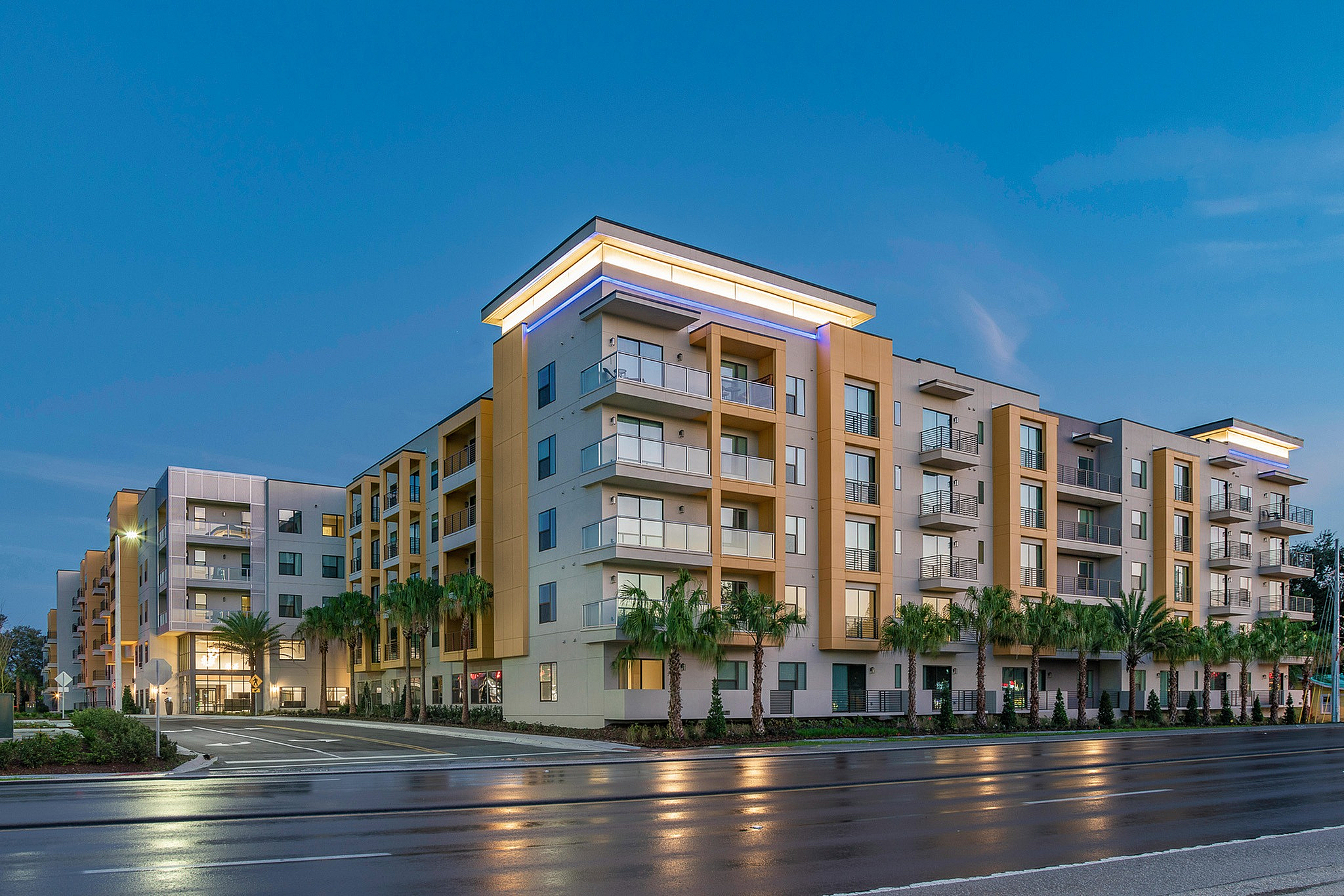 Modern luxury apartment complex at dusk, featuring sleek architecture, illuminated balconies, and lush palm trees lining the exterior.