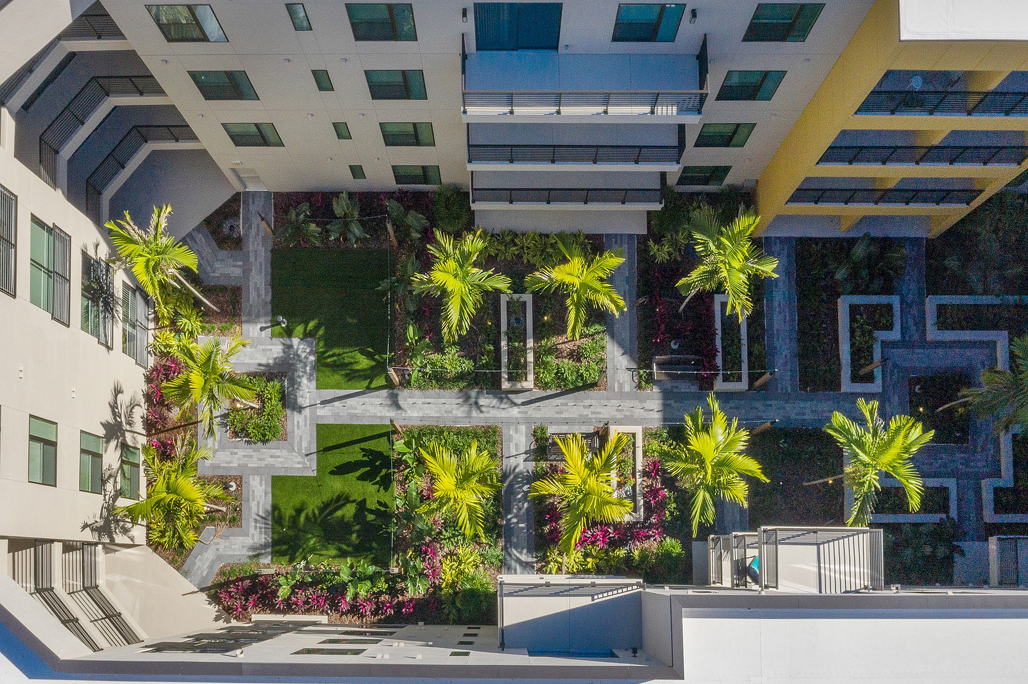 Stunning aerial view of a contemporary apartment complex courtyard, featuring lush green lawns, vibrant flower beds, and elegant palm trees.