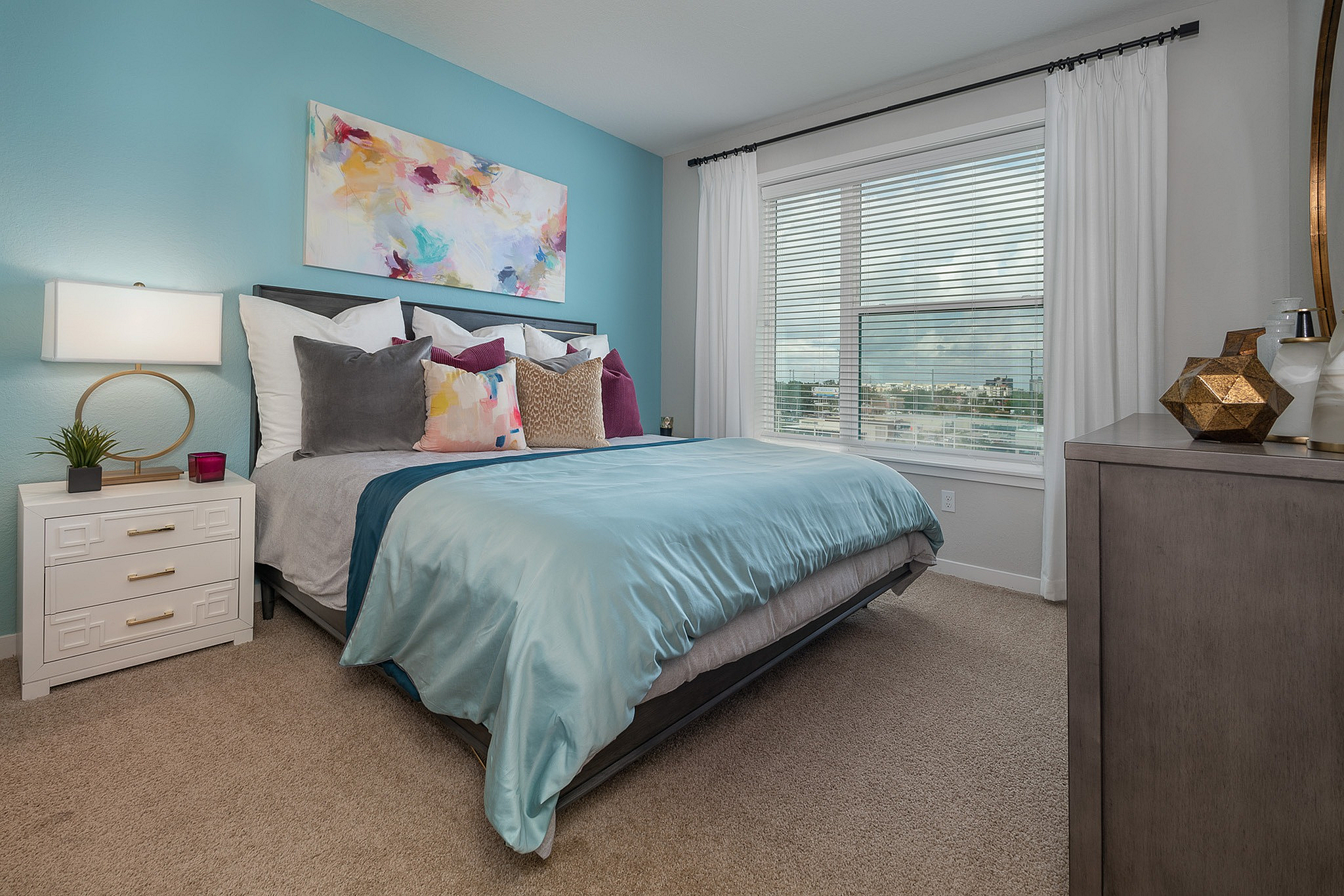 Inviting bedroom with a modern blue accent wall, cozy bed adorned with colorful pillows, large window with scenic views, and stylish decor.