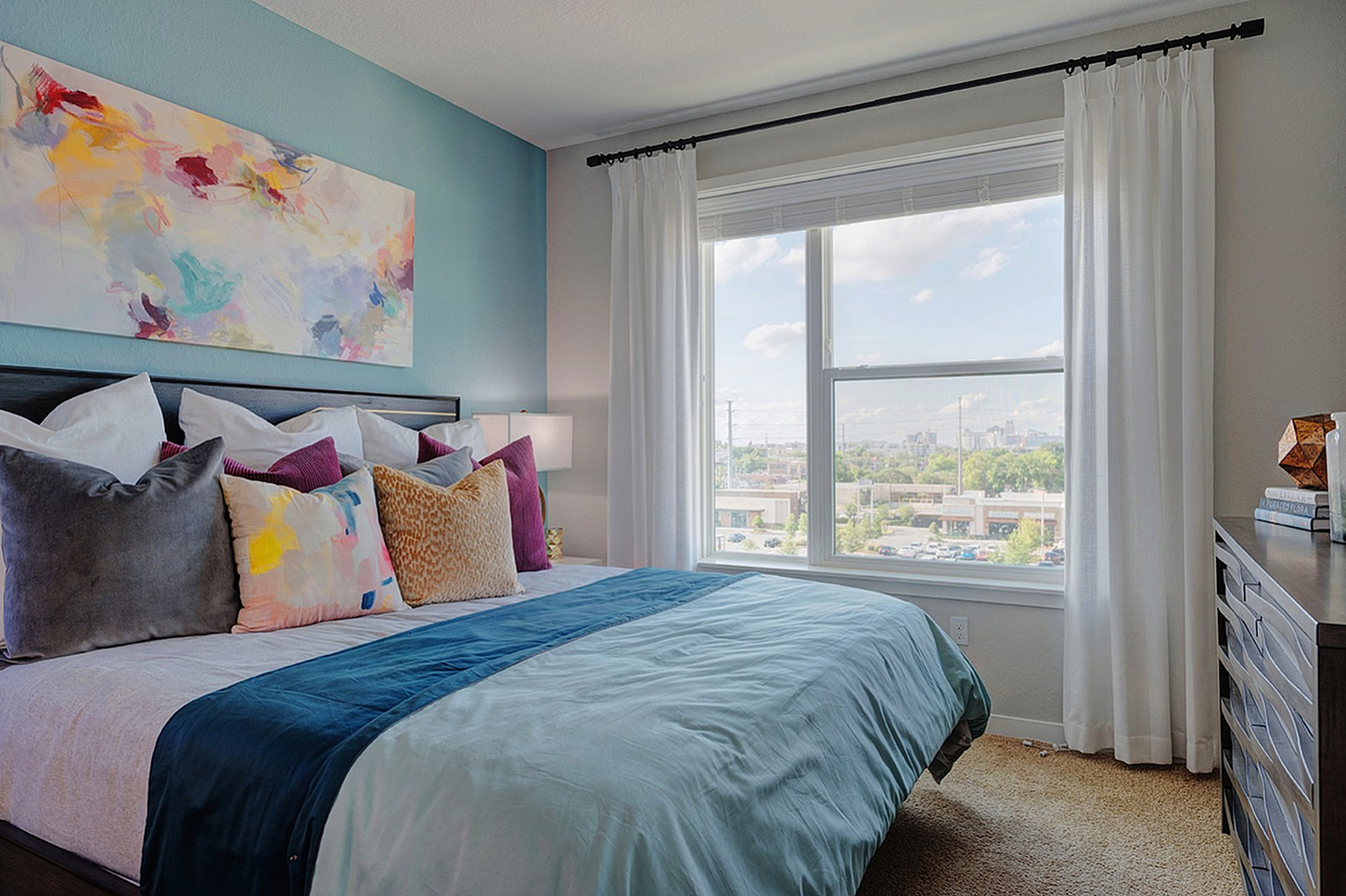 Bright bedroom with a large window view, accented by colorful abstract art and plush pillows for a cozy ambiance.