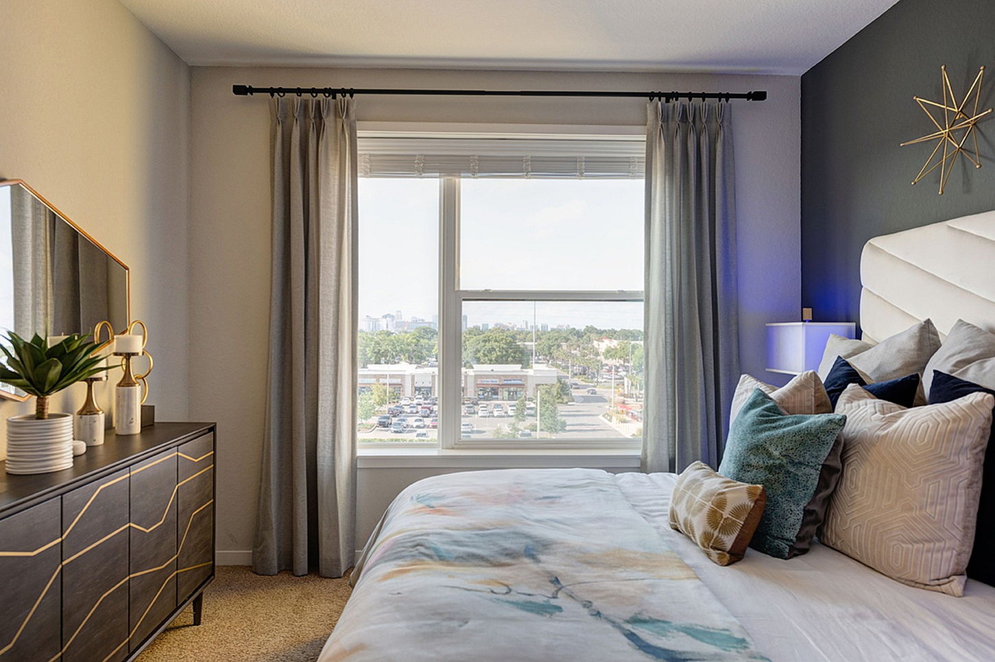Bright and cozy bedroom with elegant decor, featuring a stylish dresser, plush pillows, and a large window offering a stunning city view.