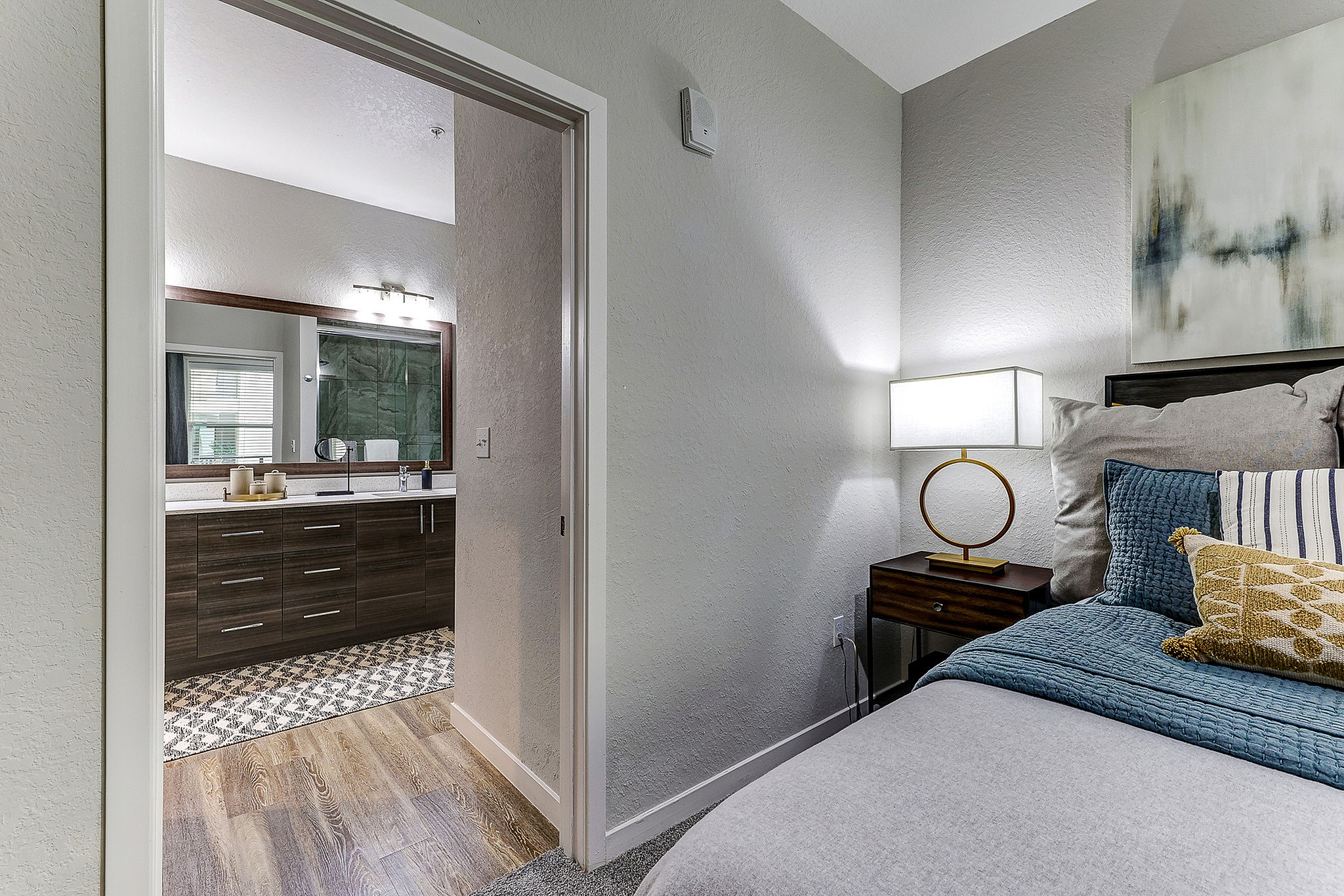Cozy bedroom with plush bedding and elegant lamp, leading into a stylish bathroom with modern cabinetry and a spacious mirror.