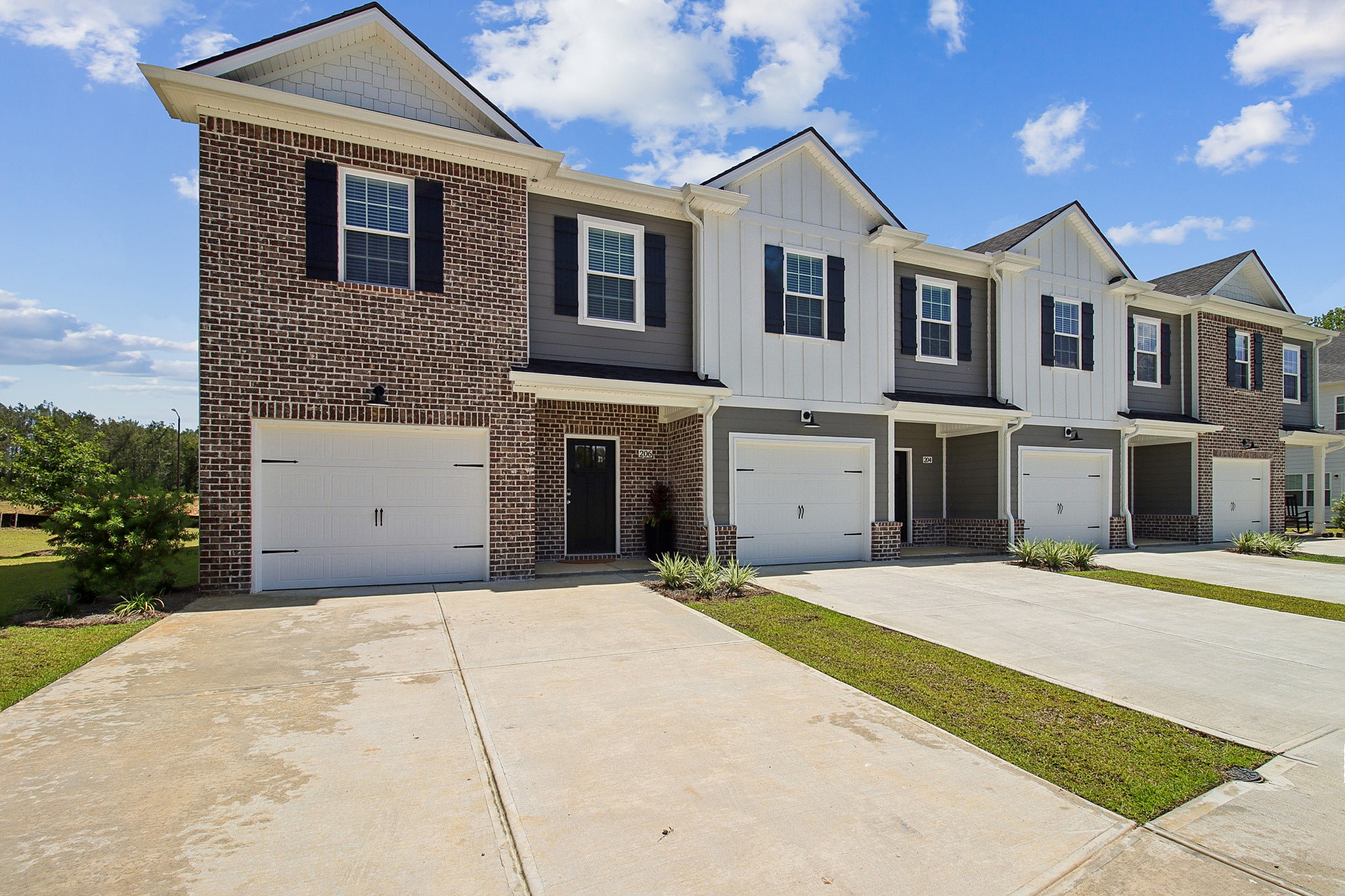 Charming modern townhouses with brick facades, featuring private driveways and lush green surroundings under a bright blue sky.