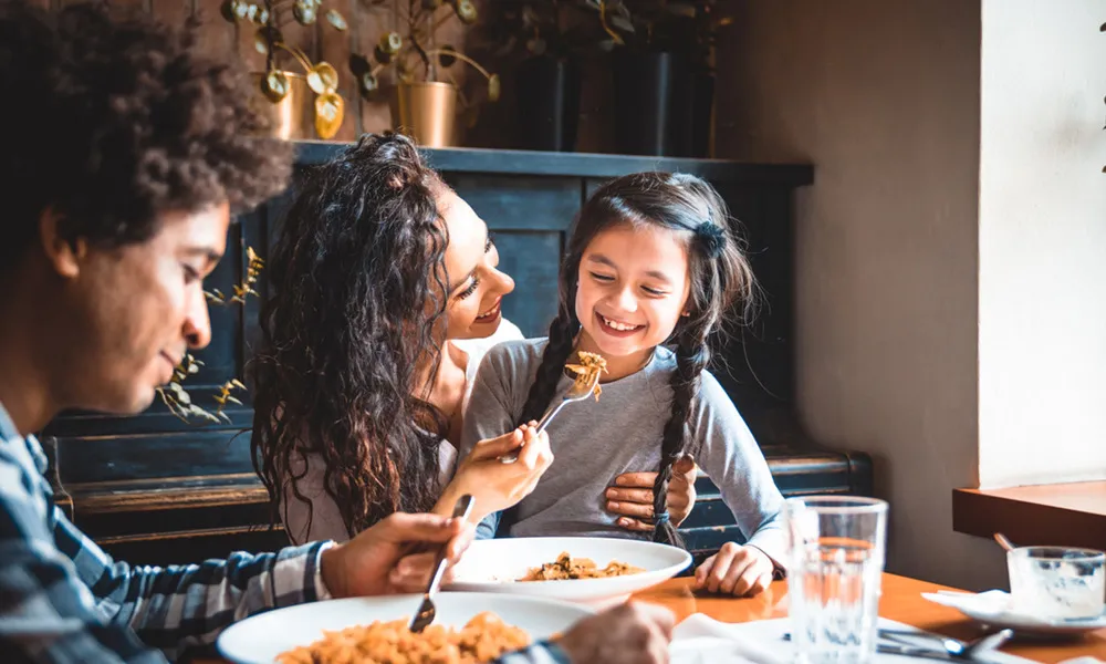 woman feeding young girl food at restaurant while man eats