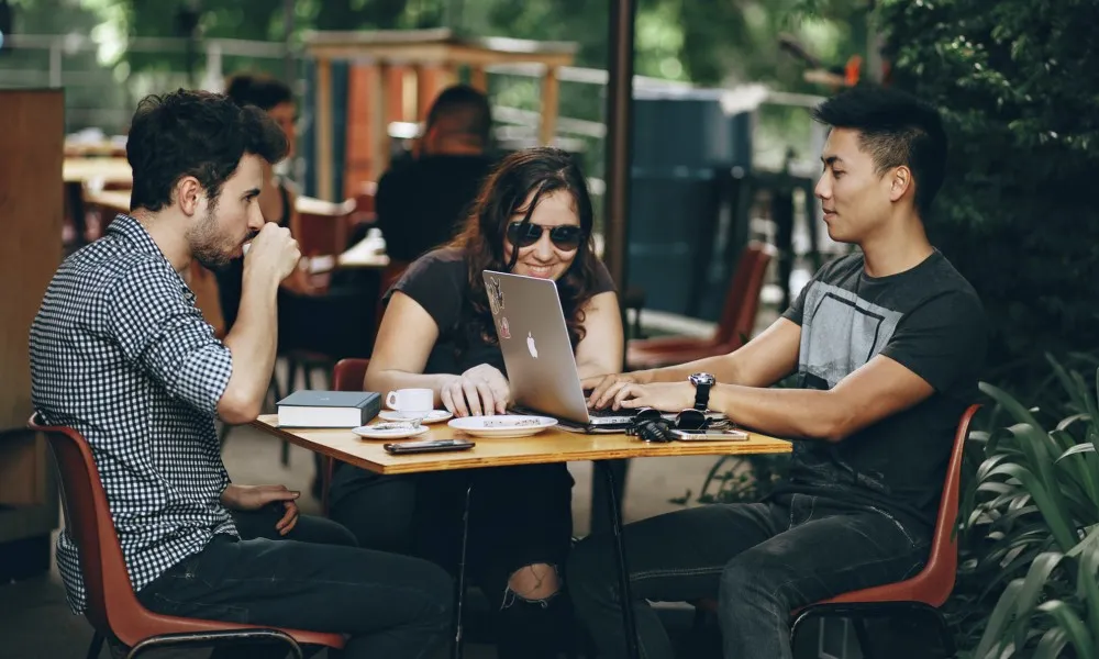 two men and a woman sitting outside of cafe with computer