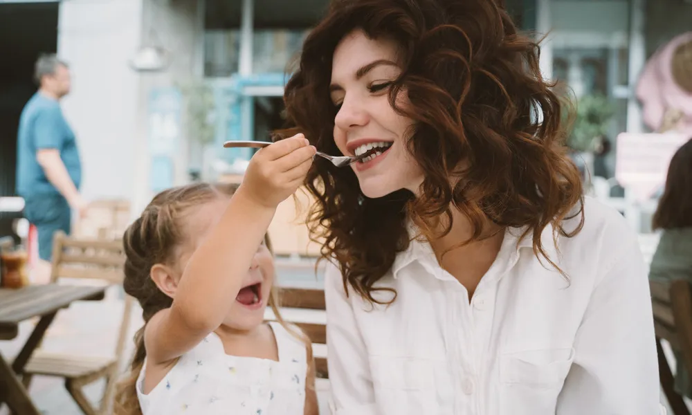 young woman being fed by child at restaurant outdoors in daytime