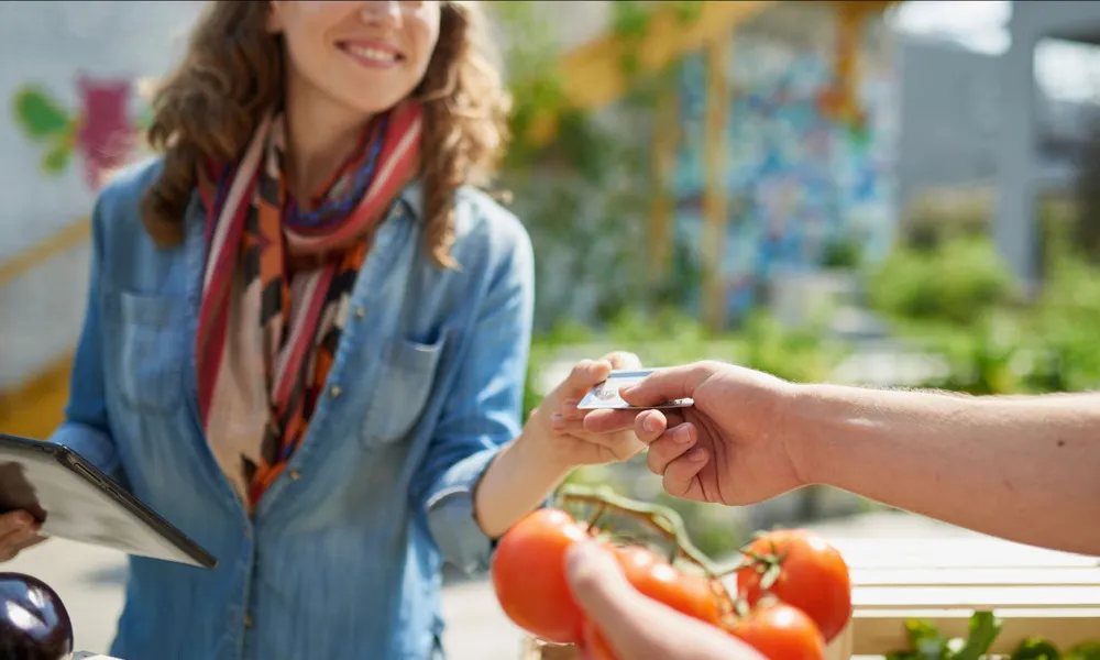 woman reaching for credit card from person holding tomatoes in outdoor market in daytime
