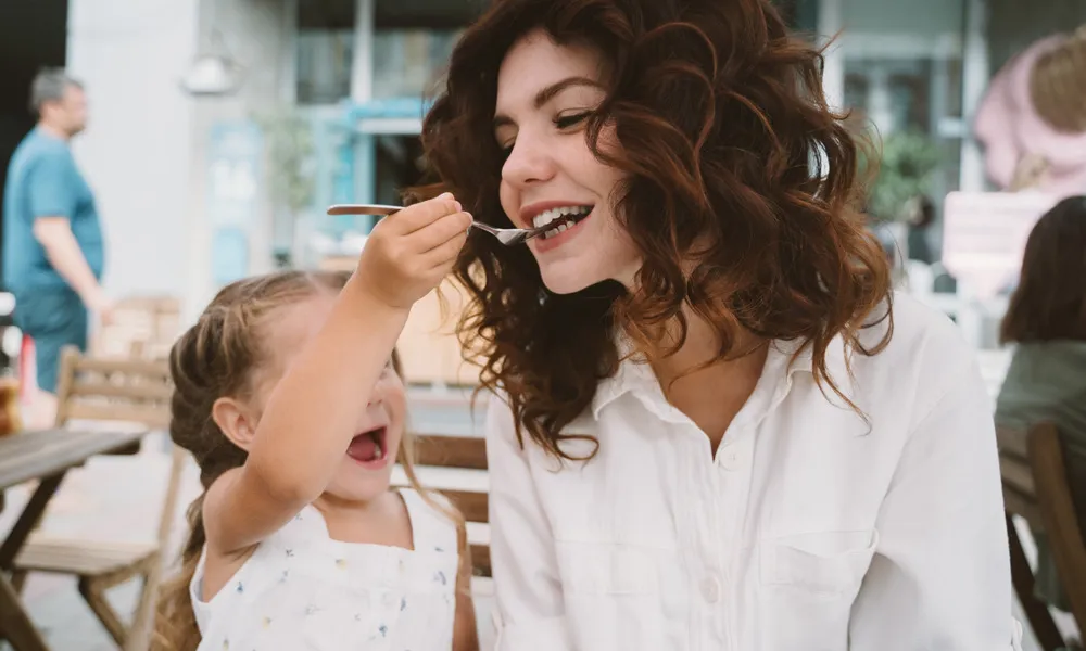 young woman being fed by small child outside in daytime at restaurant