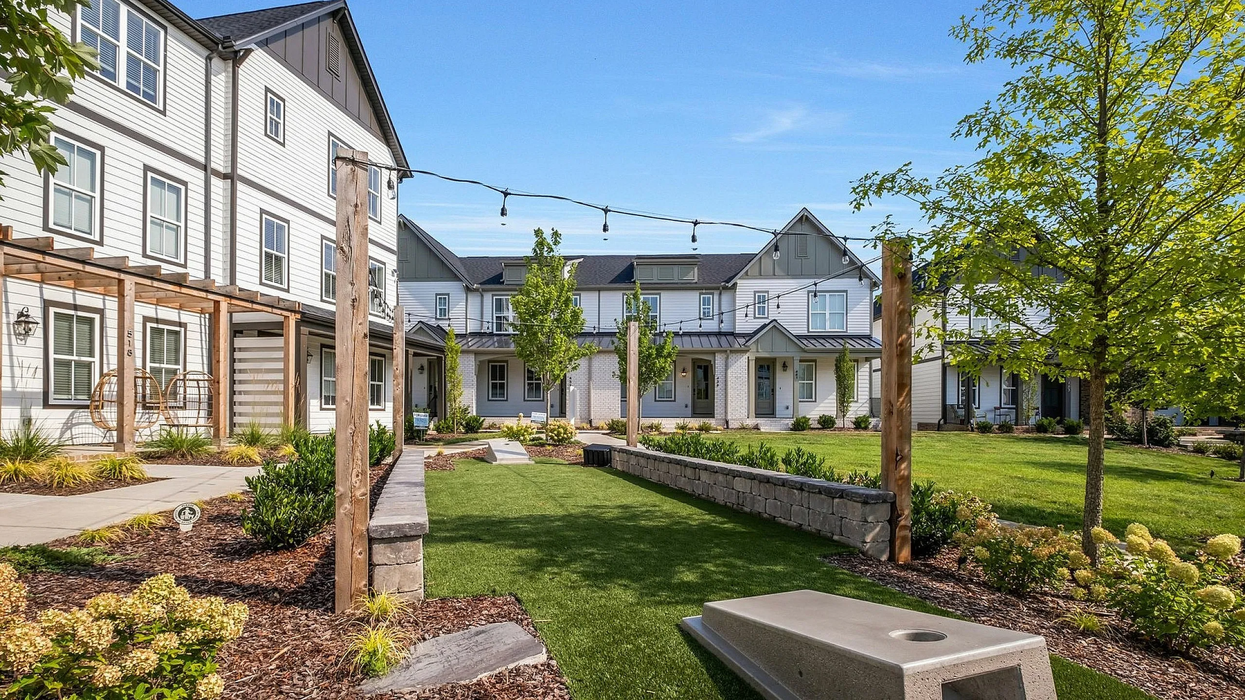 Charming outdoor space in a modern townhouse community featuring a lush green lawn, garden beds, and hanging chairs under a pergola, perfect for relaxing and socializing.