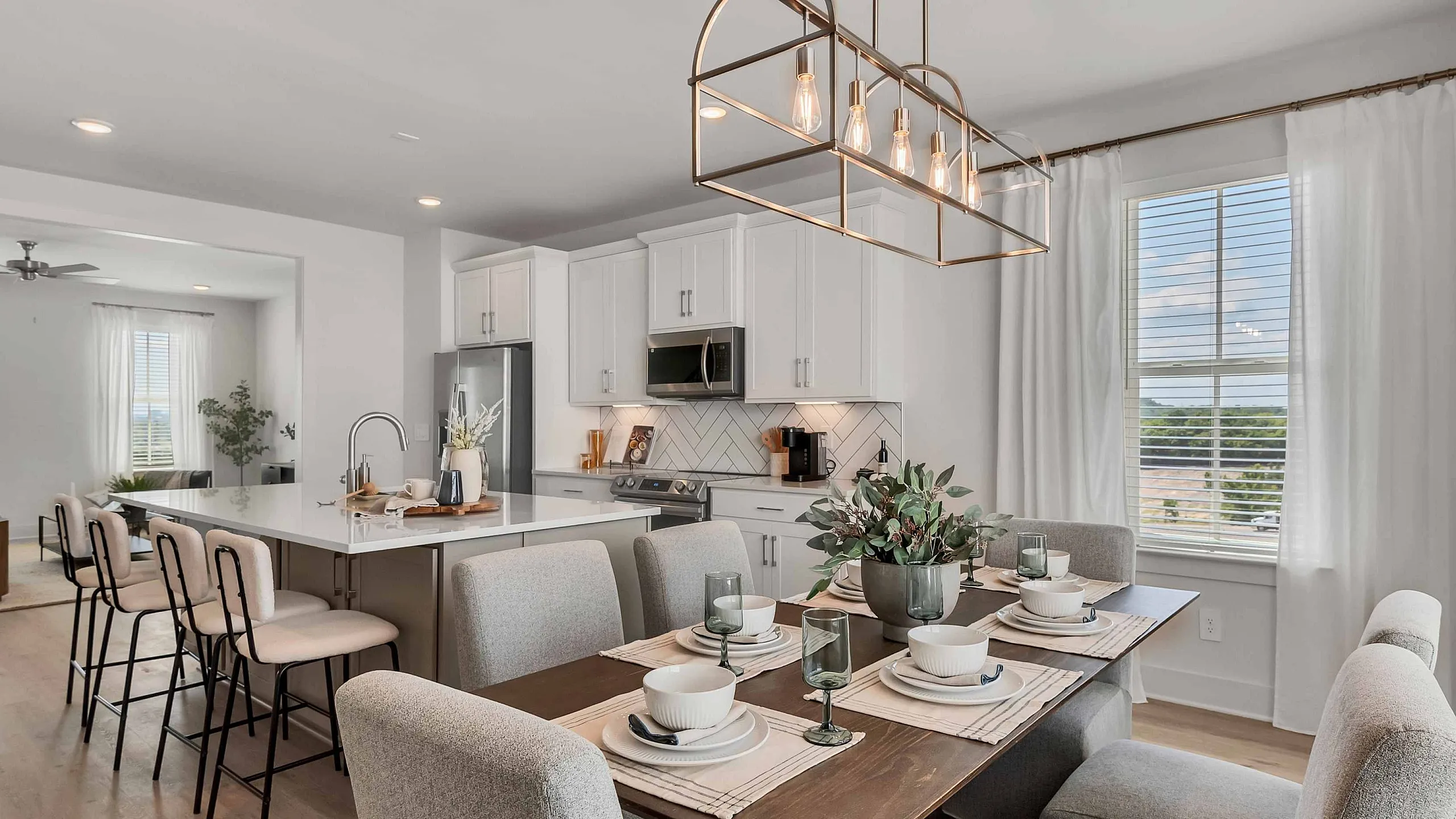 Modern open-plan kitchen and dining area with elegant decor, featuring a spacious island with bar seating, chic lighting, and a large window with natural light.