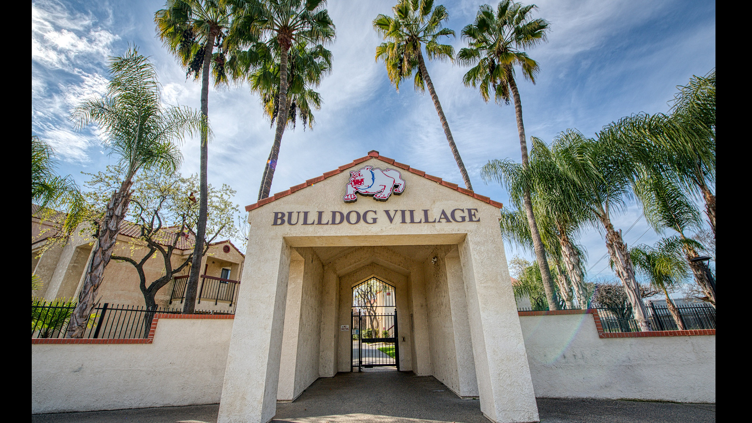 Inviting entrance to Bulldog Village with palm trees and blue sky, promising a vibrant and sunny living experience.