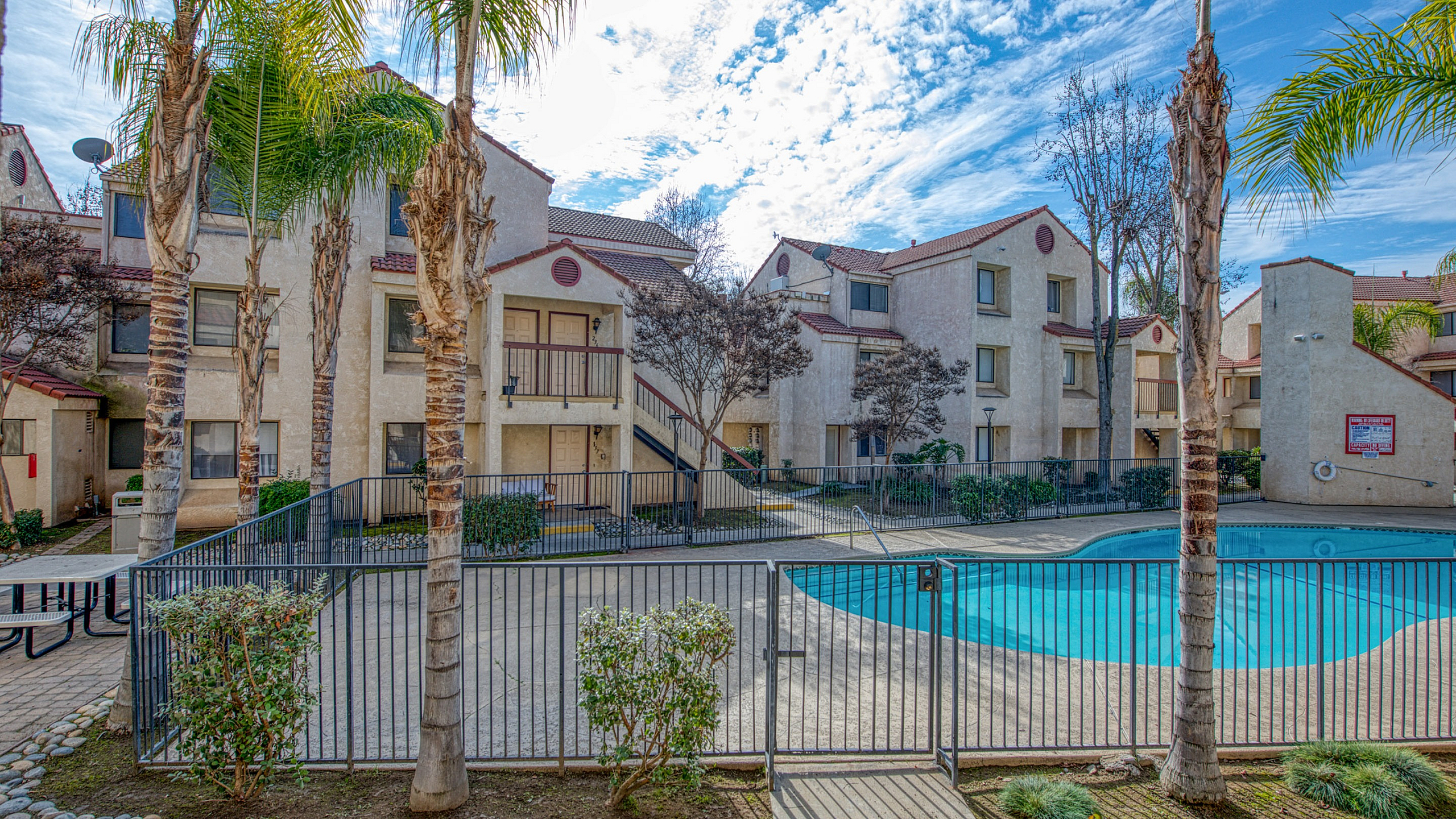 Charming apartment complex with a crystal-clear swimming pool, surrounded by lush palm trees and spacious balconies under a bright blue sky.