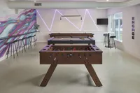 Modern community game room with foosball and pool tables, bar seating, neon wall lights, and a colorful mural.