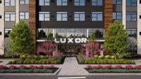 Modern apartment building entrance featuring sleek design, lush landscaping with vibrant pink blossoms, and tall green trees.