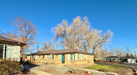 Charming single-story brick home with large windows, surrounded by mature trees and set against a bright blue sky.