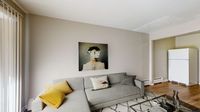 Modern living room with a stylish gray sofa adorned with vibrant yellow and patterned cushions, contemporary art on the wall, and an inviting atmosphere near the kitchen area with natural light streaming through vertical blinds.
