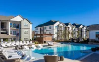 Luxurious apartment complex with a stunning outdoor pool and modern lounge area, surrounded by beautiful, multi-story residential buildings under a clear blue sky.
