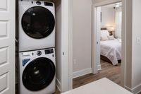 Chic apartment feature with modern stacked washer and dryer next to a cozy bedroom illuminated by natural light.