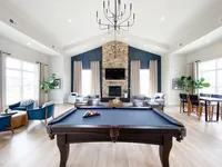 A clean, modern layout that flows from games to gathering space without feeling crowded. Spacious and elegant living room featuring a luxurious pool table, cozy seating area with a stone fireplace, high ceilings with a chandelier, and large windows allowing natural light to flood the space.