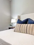 Soft touches and calming tones make this space easy to settle into. Serene bedroom corner with a cozy bed, elegant patterned cushion, and a stylish bedside lamp on a wooden nightstand.