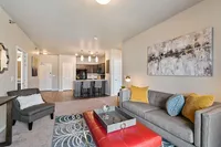 Contemporary open-concept living room with stylish decor, featuring a plush gray sofa, vibrant accent pillows, chic wall art, and a modern kitchen in the background.