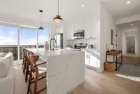 Bright modern kitchen with sleek white cabinetry, marble island, and stylish wood seating, offering panoramic city views.