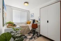 Fully furnished modern bedroom featuring a cozy bed with accent pillows, compact desk with a stylish chair, large wardrobe, and lush houseplant under a soft ceiling light.