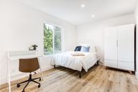 Bright and modern bedroom with large window, featuring cozy white bedding, a sleek desk with a chair, and a stylish wardrobe on elegant wood flooring.