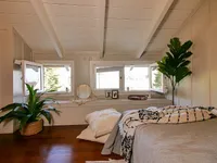 Charming attic bedroom with cozy bedding, natural light through large windows, and lush green plants for a serene ambiance.