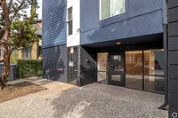 Modern apartment entrance with sleek dark blue facade, large windows, and secure entryway surrounded by greenery.
