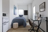 Modern small bedroom with a neatly tucked bed, a compact workspace featuring a sleek desk and chic chair, and a mini fridge microwave combo.