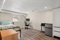 Cozy, modern bedroom with bright natural light, stylish wood flooring, compact workspace, and mini fridge.